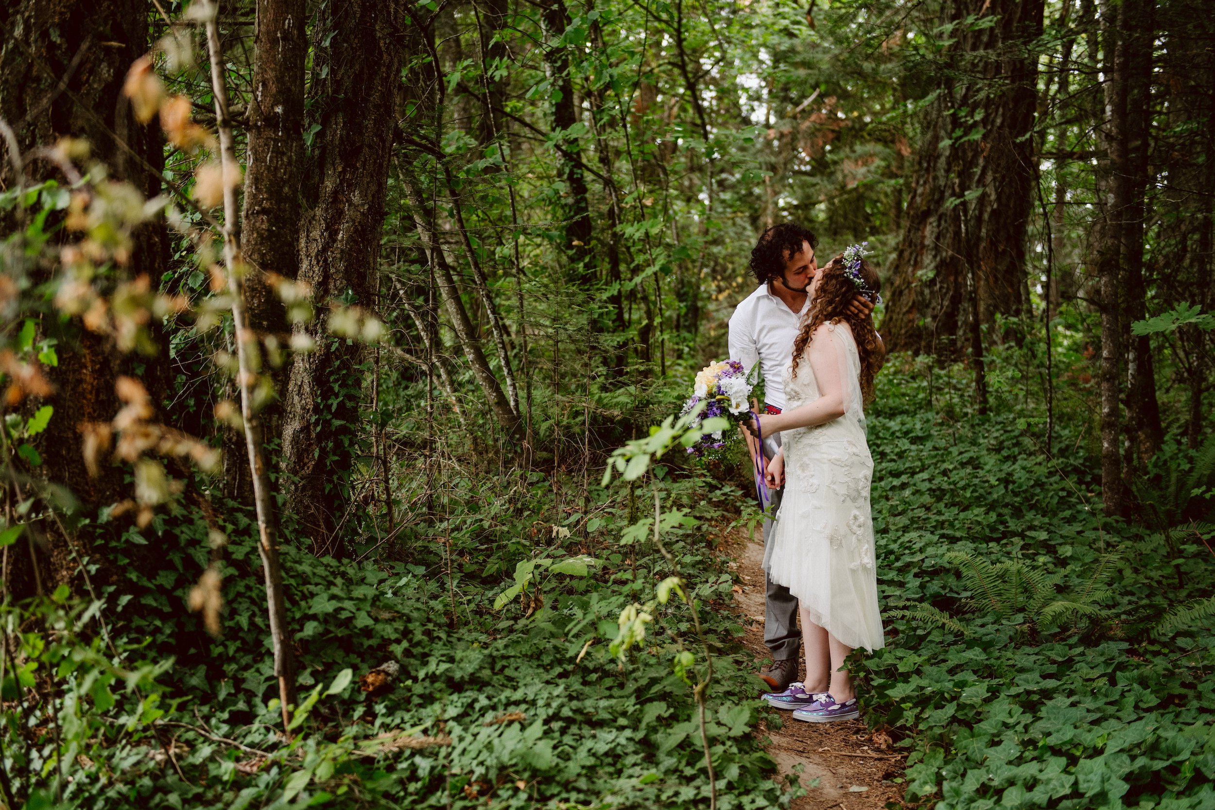 A man and a woman in wedding attire kiss in the middle of a singletrack trail in the middle of the woods