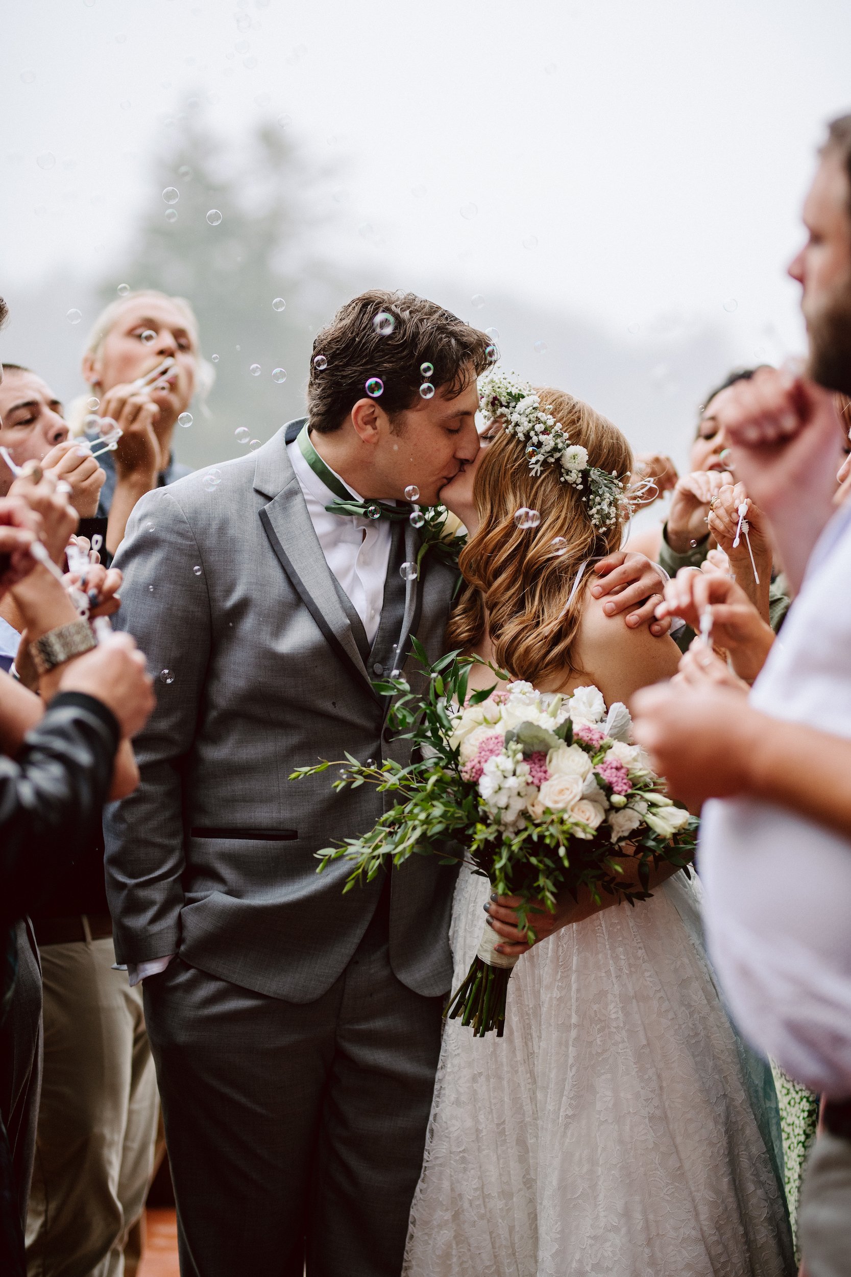 A man and woman in wedding attire kiss while their family gathers around them, blowing bubbles in celebration.