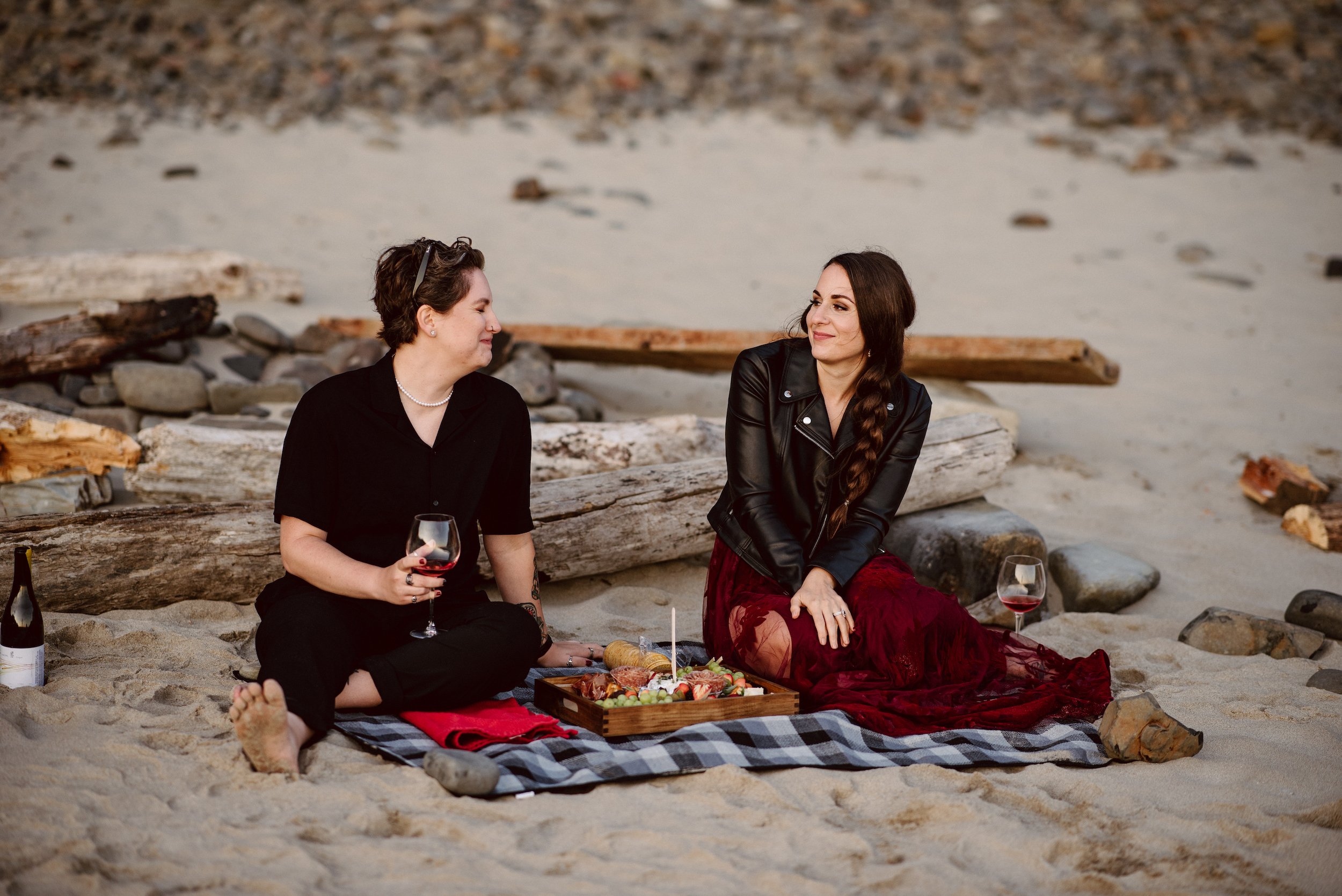 Two brides sitting on blanket on a sandy beach sharing a charcuterie board and wine and laughing together