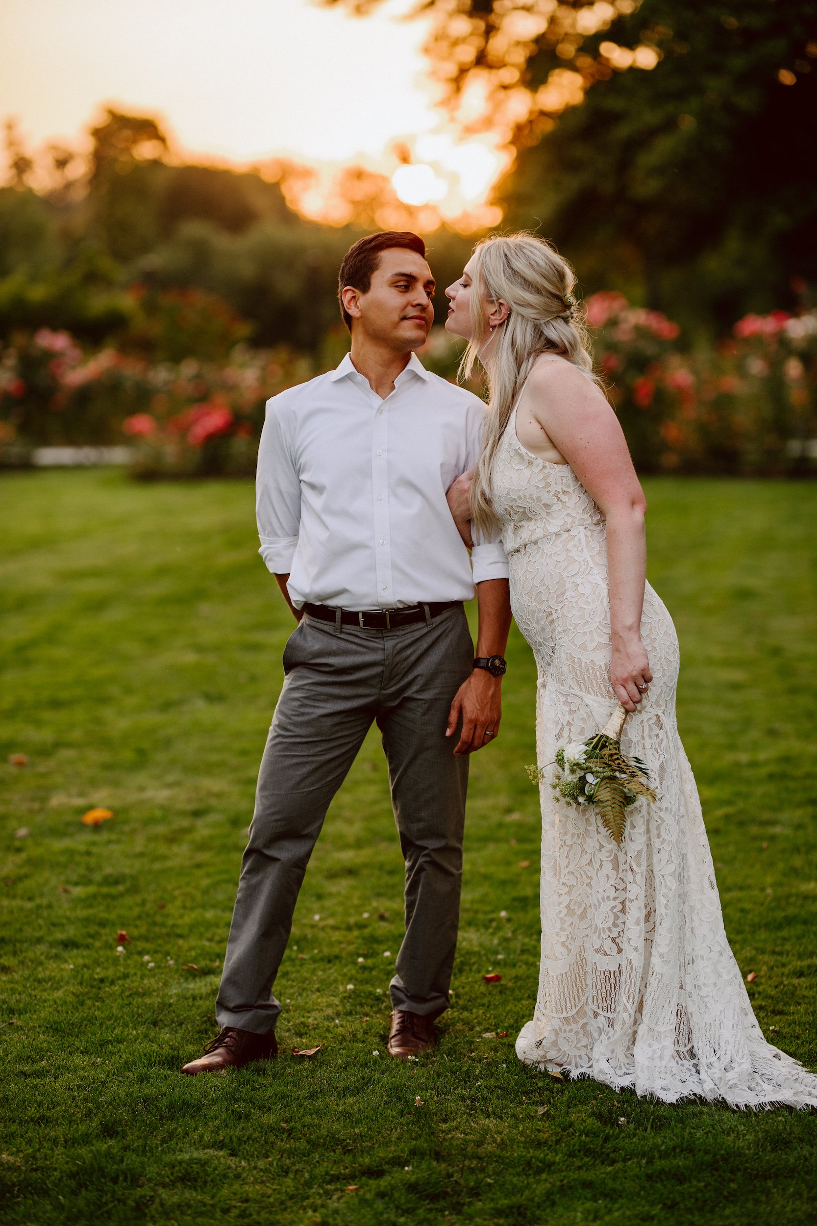 A couple in wedding attire stand together during a golden sunset in a field. They are looking at each other lovingly and standing very close to one another.