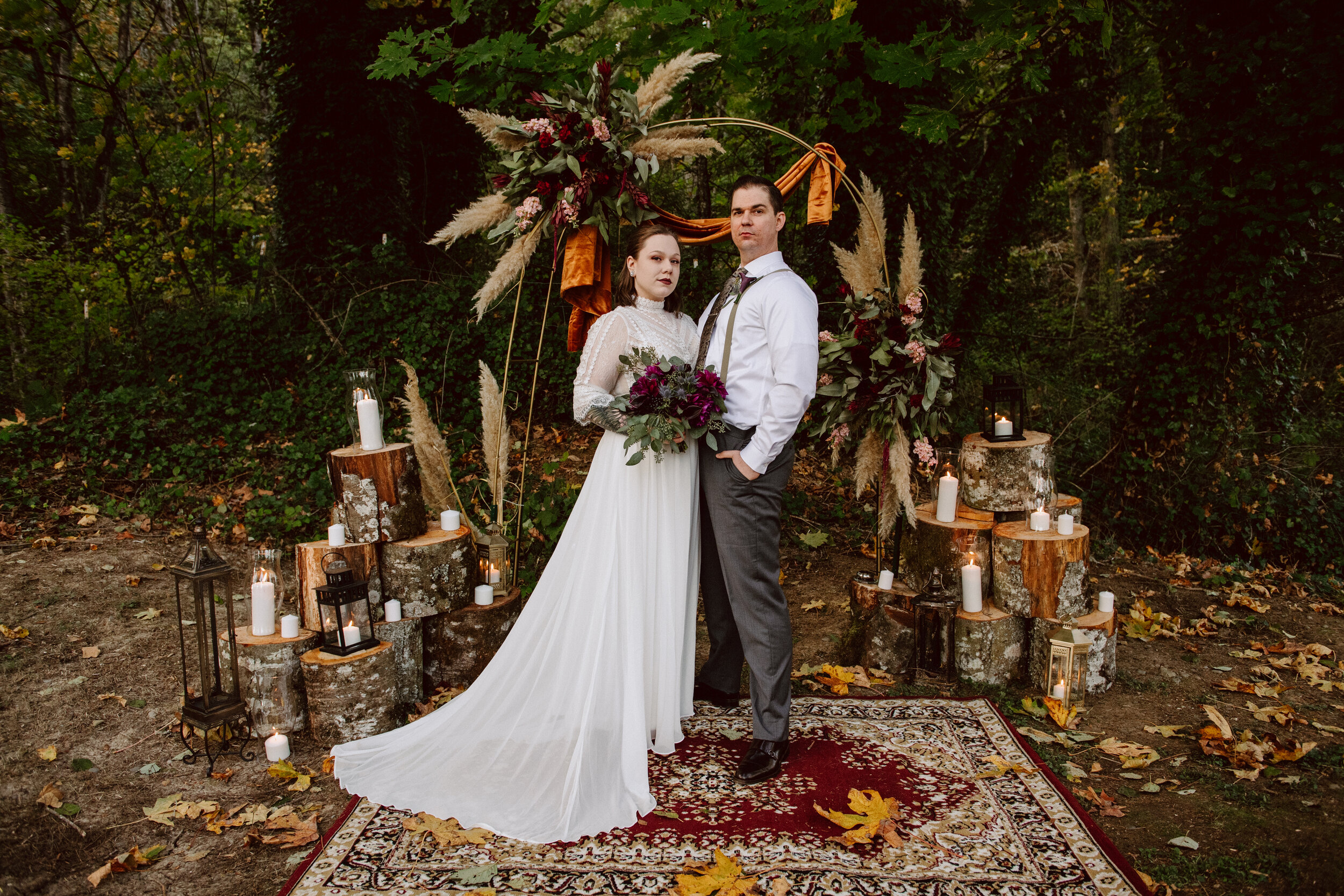 Bride in a gothic style dress holding a purple and green bouquet standing next to groom in gray pants with suspenders at their wedding altar adorned with fall florals, candles, and fallen leaves atop a vintage rug