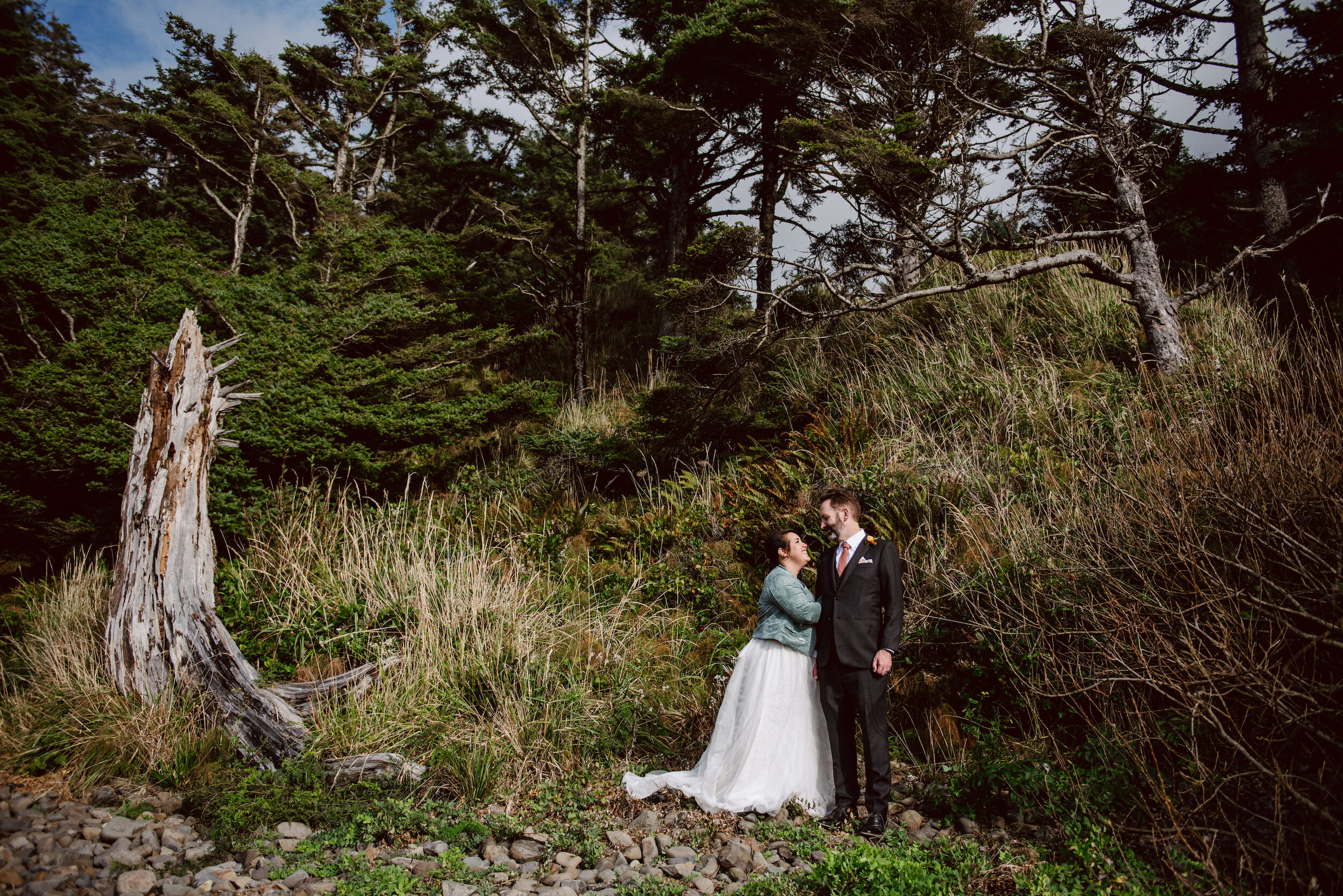 Bride in a white dress with a jean jacket cuddles the arm of a groom in a black suit while they smile at each other and stand in front of a fairytale like forest scene at the Oregon Coast