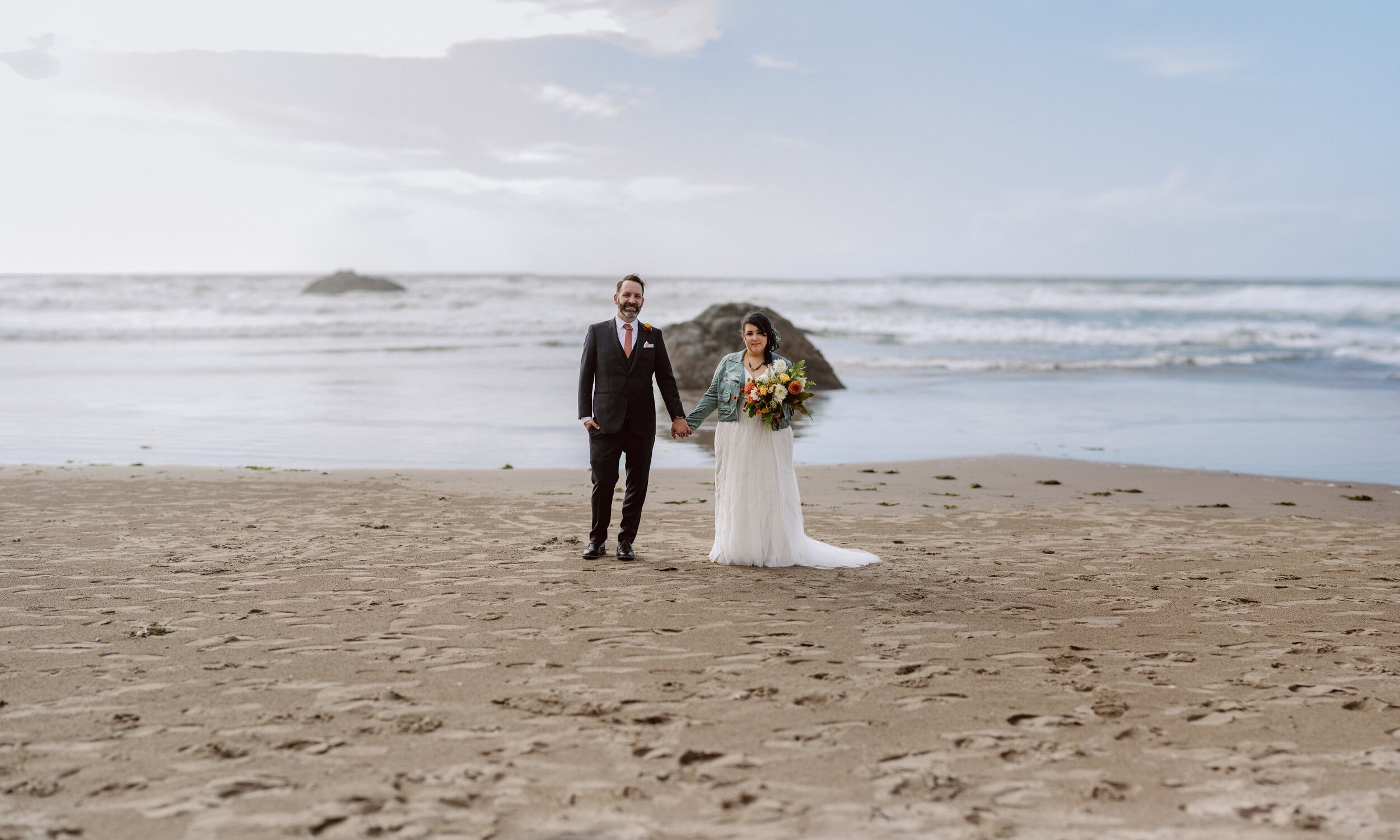 Panorama of Hug Point beach with a bride and groom in the center holding hands, the bride holding a large fall-colored bouquet