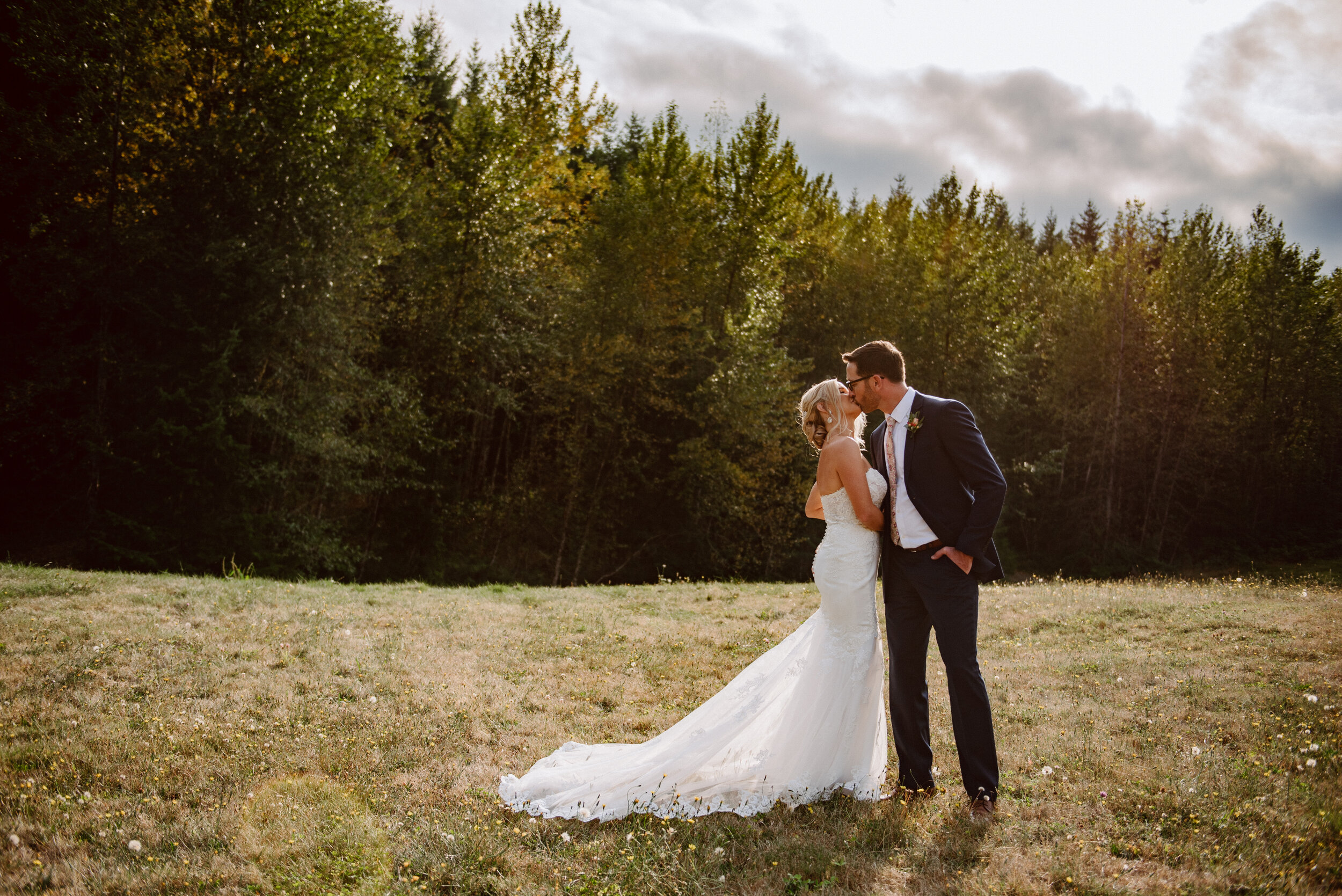 Bride and groom kiss atop a golden hill with a line of trees and stormy sky behind and the sun sets in the background