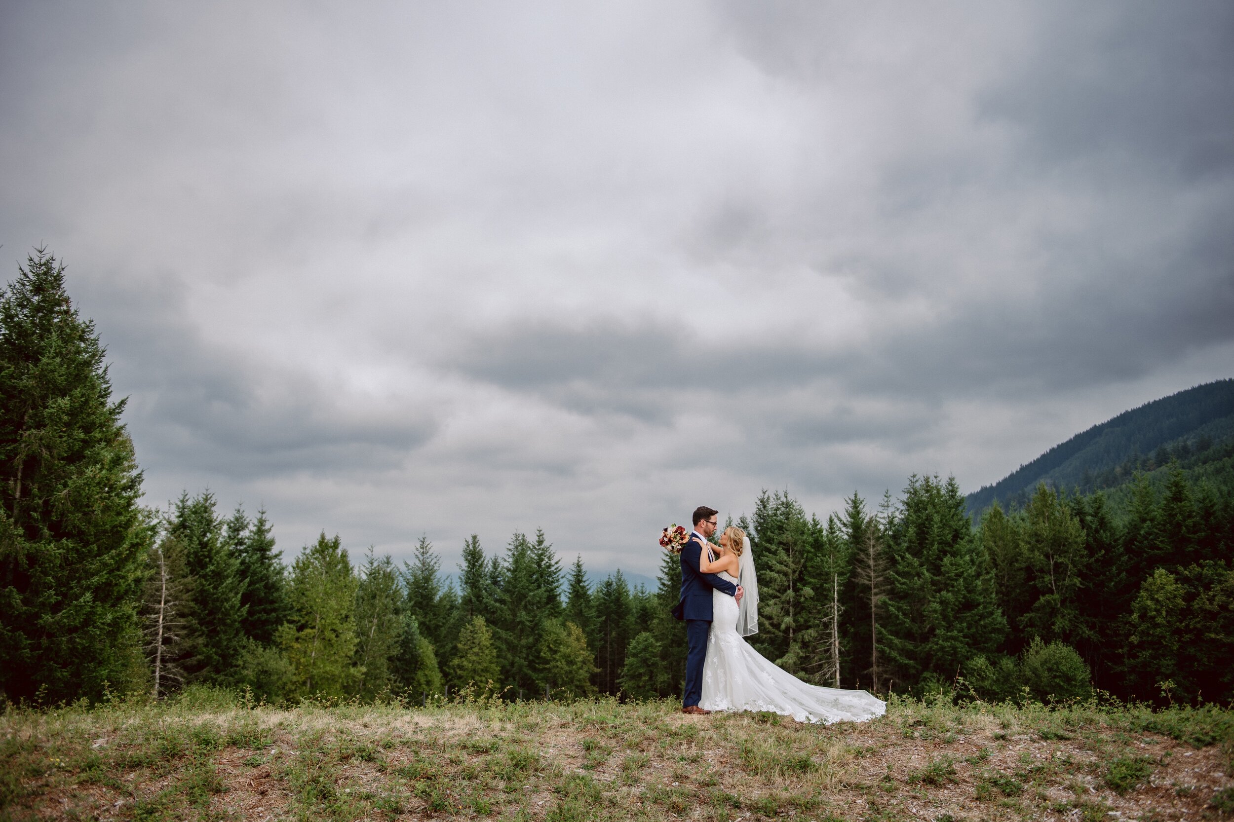 Bride and groom embrace on a hill with a forest and stormy sky behind them