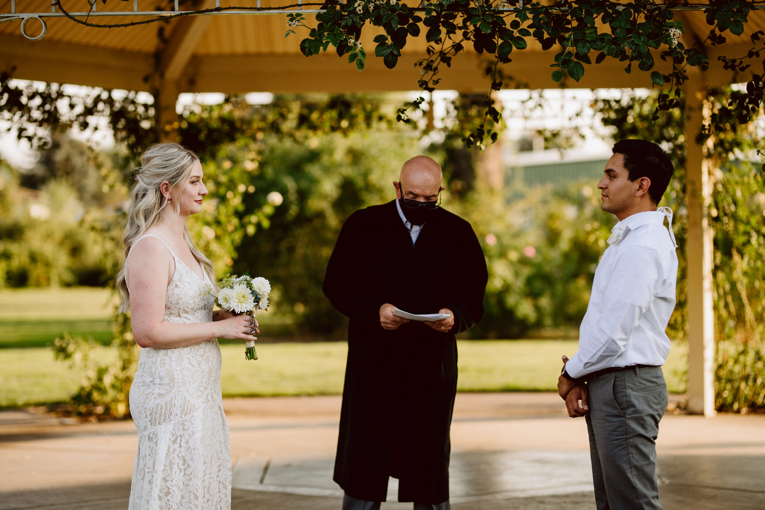 Bride and groom face each other during their elopement ceremony while an officiant reads from his notes under a gazebo at sunset in a garden