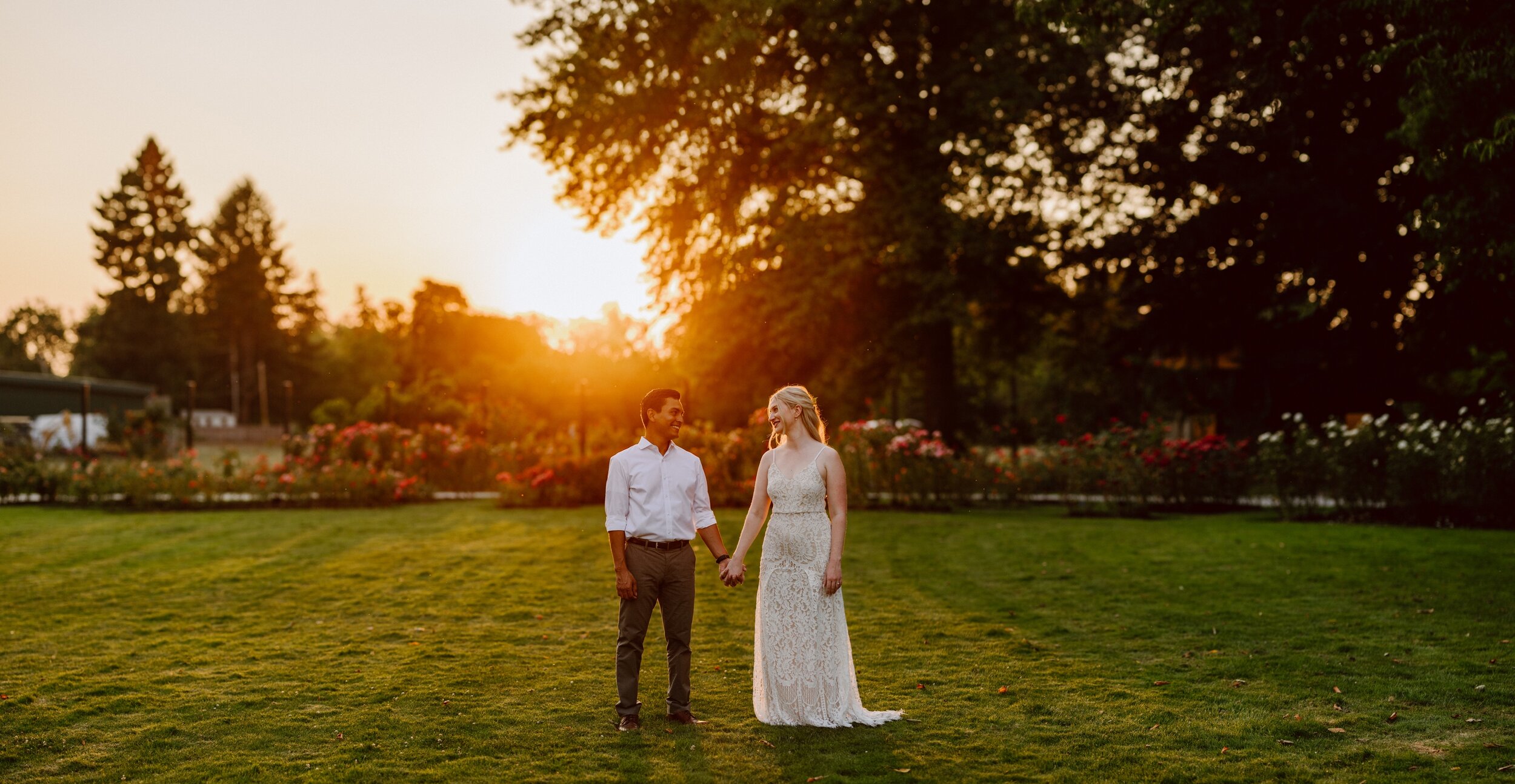 A bride and groom hold hands in a garden and smile at each other while a golden sun sets behind them