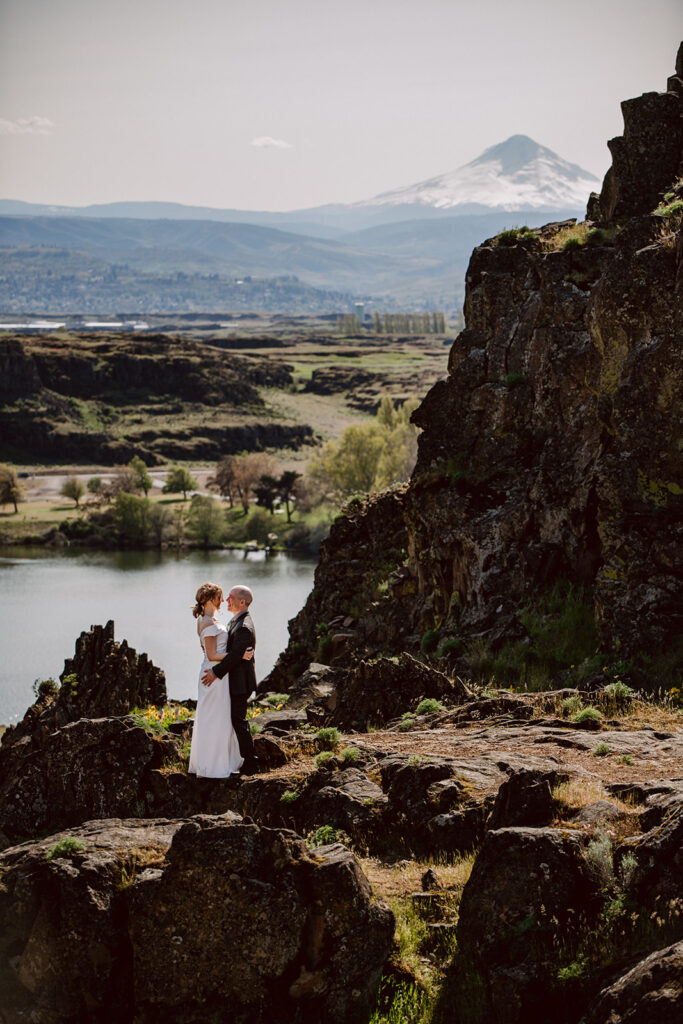 A couple in wedding attire stand atop rock formations dancing together with a the Columbia River in the background and Mount Hood in the distance