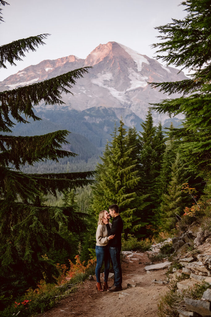 A couple playfully touches noses and embraces one another on a trail in the middle of evergreen trees with Mount Rainier, enveloped in a pink glow, in the background