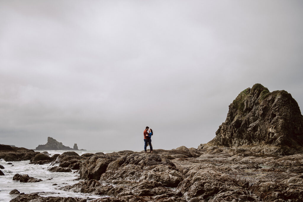 A couple stands on rock formations next to the ocean with the wind blowing against a cloudy, dark sky