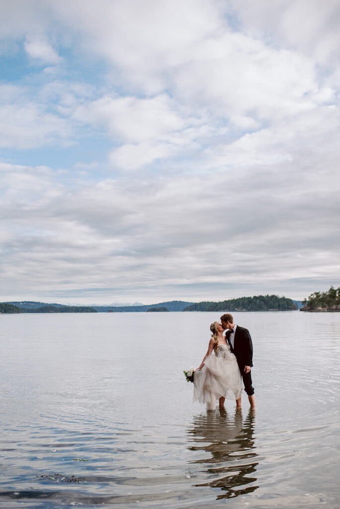 A couple in wedding attire stands in the middle of a body of water kissing and there are mountains in the far distance