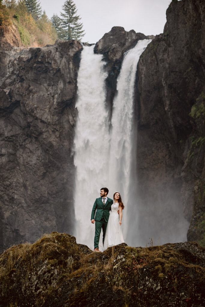 A couple in wedding attire, one in a white dress and one in a dark green suit, stand on top of a mossy rock formation with a two-columned waterfall cascading in the background behind them
